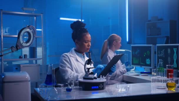アフリカ系アメリカ人女性医師がレポートをレビューし 顕微鏡下でサンプルを検査します 白い女性の研究室のアシスタントは コンピュータのキーボードを入力している 白人医療従事者又は研究者 — ストック動画
