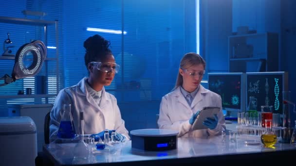 一位非洲裔美国妇女仔细检查虚拟图形 缩放并扫描图片 白色女性实验室助理将数据写入数字平板电脑 蓝光生化现代化实验室 — 图库视频影像