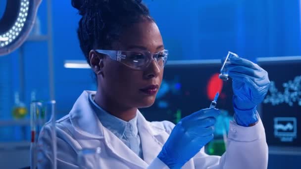 若いアフリカ系アメリカ人女性がガラス瓶から透明な液体で注射器を満たし 薬のいくつかを吐き出します 白い医療用コート ゴーグル 手袋の黒い女性医師が働いています — ストック動画