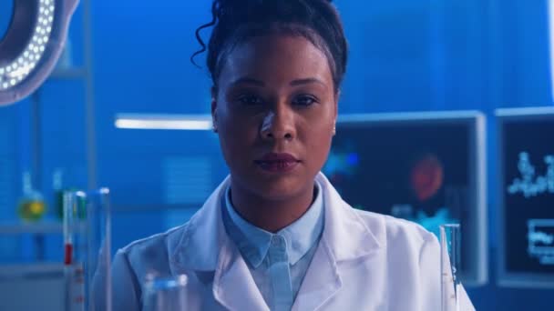 一位年轻的非洲裔美国妇女直视镜头 微笑着 一个身穿白色医疗长袍的黑人女医生穿着蓝光的现代生化实验室的画像 靠近点 — 图库视频影像