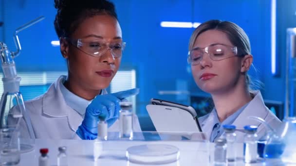 アフリカ系アメリカ人女性の医師は 棚にガラスのバイアル フラスコ 試験管の数をカウントします 白い女性研究室のアシスタントは デジタルタブレットにデータを入力します 現代研究室 — ストック動画