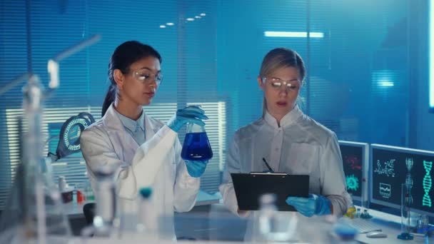 一位亚洲女医生检查了瓶中的蓝色液体样本 而一位助手则将研究数据写入了平板电脑 身穿白色医疗服的多族裔妇女 蓝色的实验室或医院 — 图库视频影像