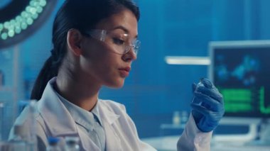 Koreli bir kadın doktor şırıngayı cam şişeden temiz bir sıvıyla dolduruyor ve ilacın bir kısmını fışkırtıyor. Beyaz önlüklü Asyalı kadın araştırmacı labaratuarda mavi ışıkla oturuyor. Biyokimyasal
