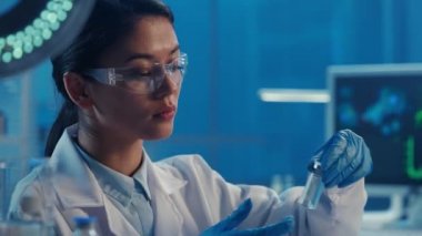 Koreli bir kadın doktor bir şişe aşı alıp onu inceliyor. Beyaz tıbbi önlüklü, gözlüklü ve eldivenli Asyalı kadın modern bir biyokimya laboratuvarında mavi ışıkla çalışıyor. Kapat.