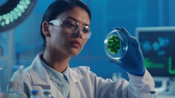 一位韩国女医生用绿色细菌样本对培养皿进行了检查 身穿白衣 戴护目镜 戴手套的亚洲女人在一个蓝光的现代生化实验室工作 — 图库视频影像