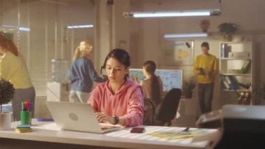 Genç bir kadın ofiste bir masada otururken kulaklıkla konuşuyor ve dizüstü bilgisayarda klavye kullanıyor. Arka planda parlak kazaklı genç yaratıcı bir yönetici takımı çalışıyor. Şey...