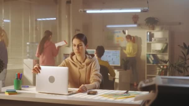 若い女性がオフィスのテーブルに座っている間 ノートパソコンのキーボードを入力しています 背景には 明るいスウェットシャツを着たマネージャーの若いクリエイティブチームが働いています オフィスワーク チームの概念 — ストック動画