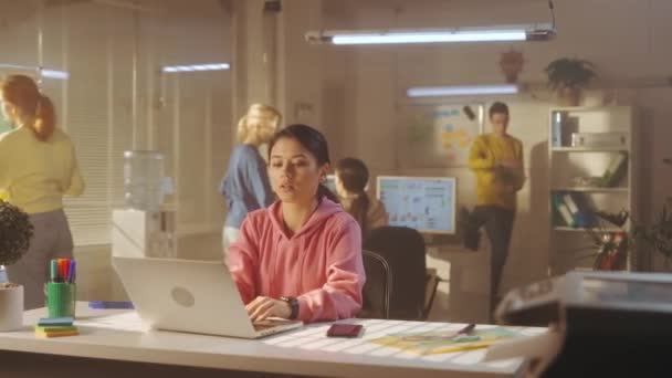 若い女性がオフィスのテーブルに座っている間 ノートパソコンのキーボードを入力しています 背景には 明るいスウェットシャツを着たマネージャーの若いクリエイティブチームが働いています オフィスワーク チームの概念 — ストック動画