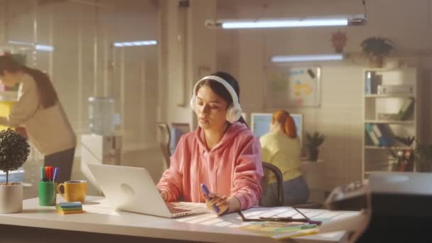 白い大きなヘッドフォンの若い女性がノートパソコンのキーボードを入力して音楽を楽しんでいます 背景には 明るいスウェットシャツを着たマネージャーの若いクリエイティブチームが働いています オフィスワークの概念 — ストック動画