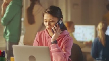 Pembe kazaklı genç bir kadın cep telefonuyla konuşuyor ve laptoptaki verileri kontrol ediyor. Arka planda, yöneticilerden oluşan genç bir yaratıcı ekip çalışıyor. Ofis işi, takım oluşturma ve...