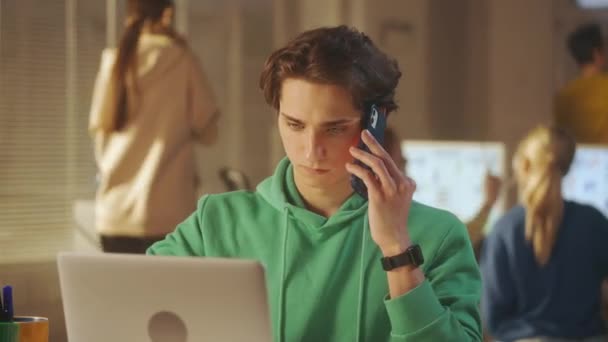 Молодой Человек Зеленой Толстовке Разговаривает Мобильному Телефону Проверяет Данные Ноутбуке — стоковое видео