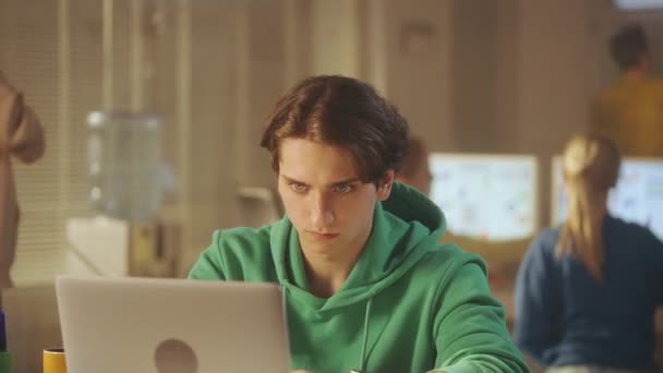 一个穿着绿色运动衫的年轻人正在笔记本电脑键盘上打字 他对自己的工作感到满意 放松下来 喝了一杯咖啡 富有创意的经理团队正在后台工作 — 图库视频影像