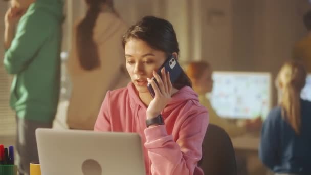 ピンクのスウェットシャツを着た若い女性が携帯電話で話し ノートパソコンでデータを確認しています 背景には 若いクリエイティブチームのマネージャーが働いています オフィスワーク チームビルディング コンセプト — ストック動画