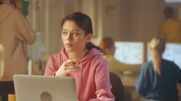 ピンクのスウェットシャツを着た若い女性がノートパソコンで働いていて オフィスのテーブルに座って何かを考えています 背景には若いクリエイティブチームが働いています — ストック動画