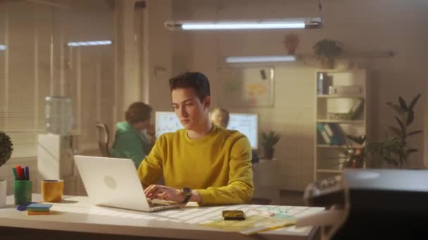 一个穿着黄色运动衫的年轻人正在笔记本电脑键盘上打字 然后放松下来 对所做的工作感到满意 一个由年轻的创意经理组成的团队正在幕后工作 办公室工作和青年 — 图库视频影像