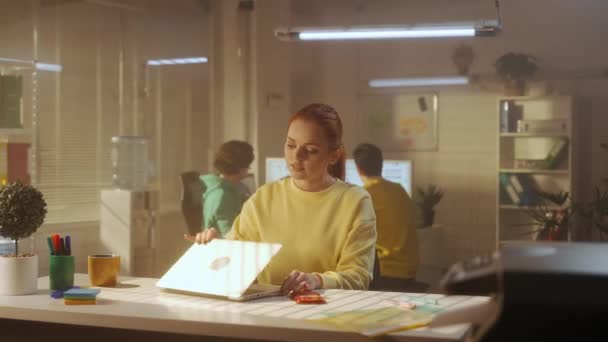 一位穿着黄色运动衫的年轻女子正在笔记本电脑键盘上打字 完成工作 摆出一副沉着的姿势放松下来 办公室里工作场所的沉思 一个由年轻的创意经理组成的团队正在工作 — 图库视频影像