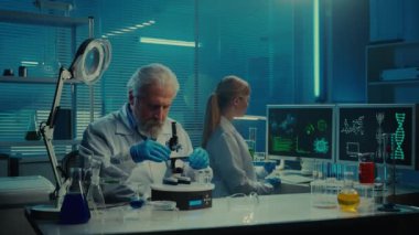 Yaşlı bir erkek doktor bir örneği mikroskop altında inceliyor. Genç bir kadın laboratuvar asistanı bir bilgisayar klavyesine yazı yazıyor ve araştırma verileri yazıyor. Beyaz önlüklü sağlık görevlileri çalışıyor.