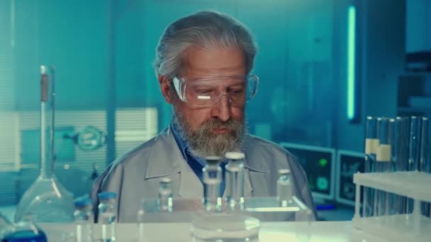 老人の医者が周りを見回すと カメラと笑顔に 白いガウンとゴーグルを着た固体の男性科学者は 青色の光を持つ研究室の背景にポーズをとります Aからの眺め — ストック動画
