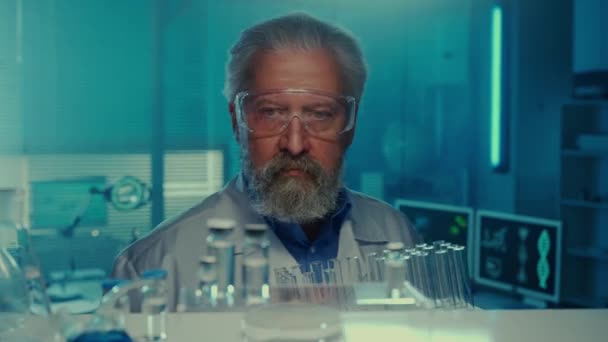 年配の男性医師が棚に試験管付きのスタンドを置きます 白いガウンとゴーグルを着た固体の男性科学者は 青色の光を持つ研究室の背景にポーズをとります 棚からの眺め — ストック動画