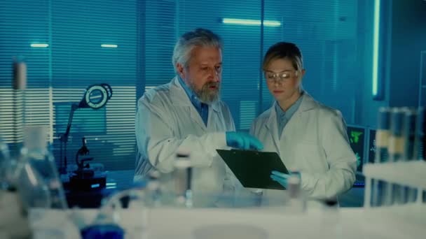 高齢の男性医師は在庫や医薬品を注文する 若い女性アシスタントはフォルダにデータを書き込みます 青い光の研究室で白衣を着た男性科学者 Aからの眺め — ストック動画