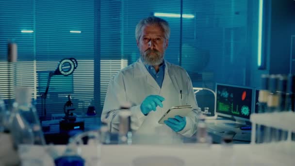 老年男医生采集库存或订购药品 并将数据输入数字平板电脑 身穿白色医疗外套 身披蓝光的坚实的男性科学家 从架子上看 — 图库视频影像