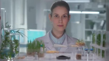 Beyaz önlüklü kadın araştırmacı kameraya bakıp gülümsüyor. Bitkilerin matara ve test tüplerinde yetişen yeşil filizleri. Laboratuvarda yetişen yeşil lahanalarla biyoloji deneyi, eğitim bilimi