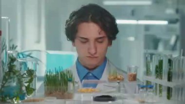 Beyaz önlüklü bir biyoteknoloji uzmanı kameraya bakıp gülümsüyor. Bitkilerin matara ve test tüplerinde yetişen yeşil filizleri. Laboratuvarda yetişen yeşil lahanalarla biyoloji deneyi. Biyoteknoloji.