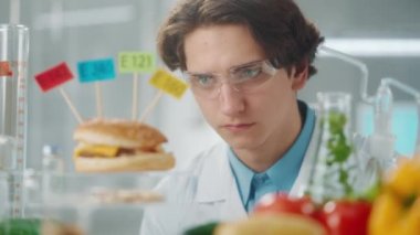 Genç erkek araştırmacı E142, E240, E121 ve E160 etiketli bir hamburger örneğini inceliyor. Biolog örneğin kalitesini analiz eder. Biyoloji deneyi Biolab 'da gıda ve sebzelerle yapılıyor. Eko ürünler, GDO