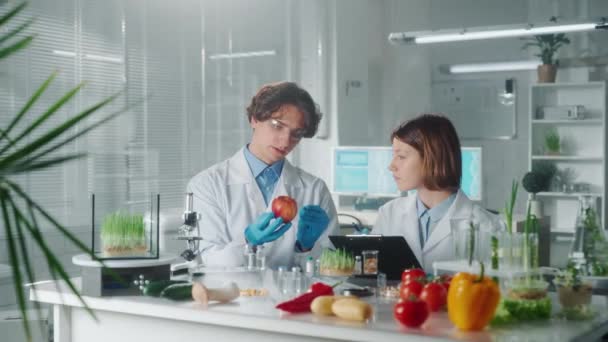 Manlig Forskare Undersöker Ett Rött Äpple Medan Kvinnlig Laboratorieassistent Skriver — Stockvideo