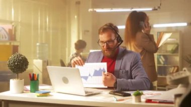 Orta yaşlı bir adam bilgisayar ve kulaklık kullanarak video konferans aracılığıyla iletişim kuruyor. Mavi ceketli ve kırmızı balıkçı yakalı bir işadamı ofisteki bir masada oturup grafik ve çizelgeleri gösteriyor.