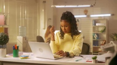 Genç bir Afrikalı Amerikalı kadın dizüstü bilgisayarda klavye kullanıyor, görme sorunu yaşıyor ve gözlük takıyor. Sarı kazaklı siyah bir kadın ofisteki bir masada oturuyor. Yönetim.