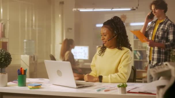 一名年轻的非洲裔美国妇女使用笔记本电脑和耳机通过视频会议进行交流 黑人女性程序员或设计师坐在办公室的桌子旁 展示图表 — 图库视频影像
