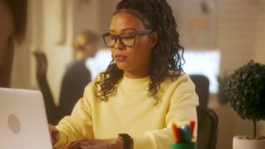 Genç bir Afrikalı Amerikalı kadın dizüstü bilgisayarda klavye kullanıyor, kameraya bakıyor ve gülümsüyor. Sarı kazaklı siyah bir kadın ofisteki bir masada oturuyor. Yönetim ekibi burada çalışıyor.
