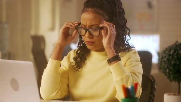 アフリカ系アメリカ人の若い女性がノートパソコンのキーボードを入力し 頭痛に苦しんでいます 黄色のスウェットシャツを着た黒人女性がオフィスのテーブルに座っている マネジメントチームは — ストック動画