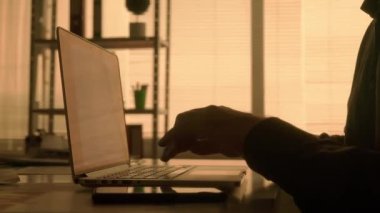 Laptop klavyesine yazı yazan ve cep telefonuyla mesajlaşan bir adamın karanlık görüntüsü. Siyah gömlekli bir erkek yönetici, tasarımcı veya programcı karanlıkta bir masada otururken çalışır.