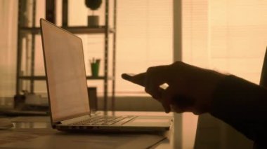 Laptop klavyesine yazı yazan ve cep telefonuyla mesajlaşan bir adamın karanlık görüntüsü. Siyah gömlekli bir erkek yönetici, tasarımcı veya programcı karanlıkta bir masada otururken çalışır.