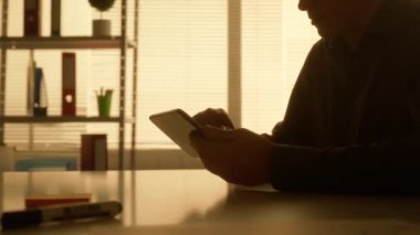 Bir adamın karanlık silueti dijital tabletinin ekranına bilgi aktarır ve yakınlaştırır. Siyah gömlekli bir erkek yönetici, tasarımcı veya programcı karanlıkta bir masada otururken çalışır.