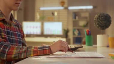 Hesap makinesine güvenen ve verileri rapora yazan genç bir kadının yan görüntüsü. Bir kadın yönetici, tasarımcı ya da programcı bir kalem ile bir kağıt parçasına bilgi yazar. Ofis işi