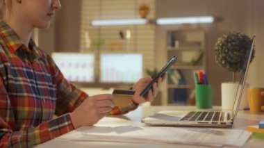 Banka kartı ve cep telefonu kullanarak online alışveriş yapan genç bir kadının yan görüntüsü. Renkli kareli gömlekli bir kadın yönetici, tasarımcı ya da programcı. Ofis işi, serbest çalışma, eğitim.