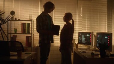 Bir erkekle bir kadının siluetleri karanlık bir ofiste yeni bir projeyi ya da dijital tabletle ilgili bir raporu tartışıyorlar. Yöneticiler büyük bir pencerenin yanında dururken iletişim kurarlar.