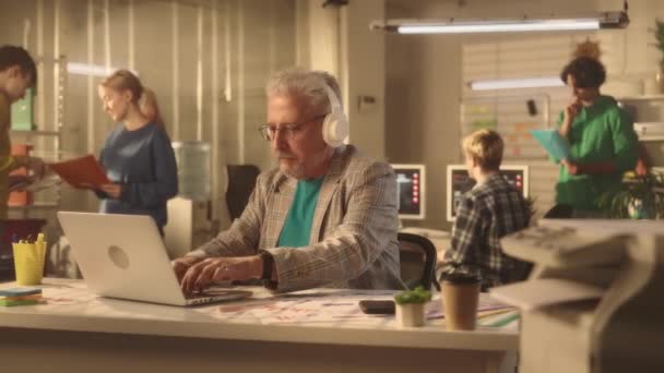 老人在笔记本电脑键盘上打字 喜欢用白色大耳机听音乐 一个商人坐在一张桌子旁的办公室里工作 一个由年轻的经理组成的富有创意的团队正在 — 图库视频影像