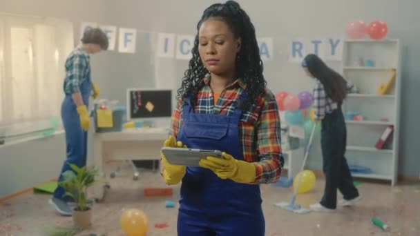 非洲裔美国女青年将办公室清洁数据输入数字平板电脑 一群身穿蓝色工作服的跨国洗衣工正在清扫地板 并在聚会后捡垃圾 规划工作 — 图库视频影像