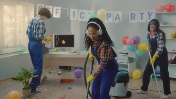 パーティーの後にオフィスを掃除する多国籍グループ アフリカ系アメリカ人の女性アジア系の女性とヨーロッパ系の男性が床を掃き掃除機でゴミ袋に入れて踊り — ストック動画
