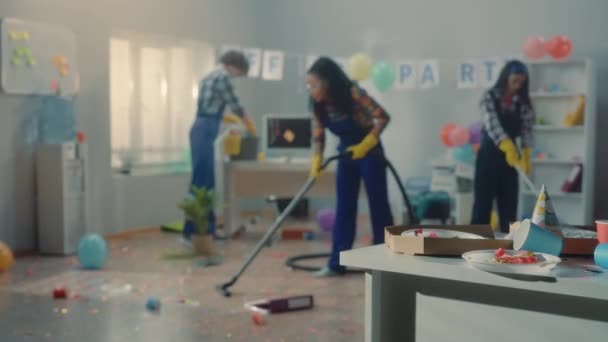 クリーナーのグループは 企業のパーティーの後にオフィスをクリーンアップします アフリカ系アメリカ人の女性 アジア系の女性 ヨーロッパ系の男性が床を掃き出し 掃除機でゴミ袋に入れて回収する 焦点は — ストック動画
