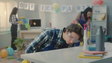 Genç bir erkek temizlikçi masaya sprey sıkar ve tozu siler. İşçi masayı bir bezle siler ve temizliği bir parmakla kontrol eder. Mavi tulumlu temizlikçiler çöpleri temizler ve...