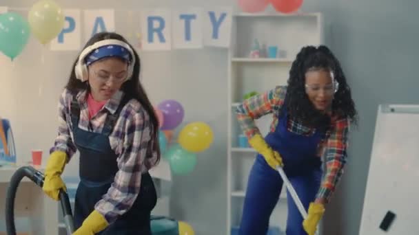 パーティーの後にオフィスを掃除する多国籍グループ アフリカ系アメリカ人の女性とアジア系の女性が床を覆い 真空とダンスを元気に音楽に合わせています 清掃サービスの概念 — ストック動画