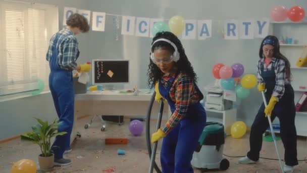 清掃員のグループはパーティー後にオフィスを掃除します アフリカ系アメリカ人の女性 アジア系の女性 ヨーロッパ人の男性が床を掃き掃除機でゴミ袋にゴミを集め 音楽に合わせて元気に踊る — ストック動画