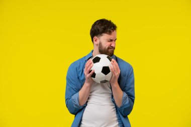 Beyaz tişörtlü heyecanlı genç sakallı adam futbol fanatiği sarı arka planda izole edilmiş futbol topu gibi bağıran favori takımı neşelendiriyor. İnsanlar aile eğlencesi konseptini kullanır..