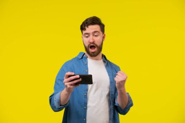 Aşırı neşeli, bronzlaşmış, mavi, basit tişörtlü yakışıklı adam telefon pozu vererek yumruğunu kaldır turuncu sarı stüdyo arka planında. Uzayın afiş modelini kopyala. Çevrimiçi sosyal medya. Şanslı kazanma konsepti