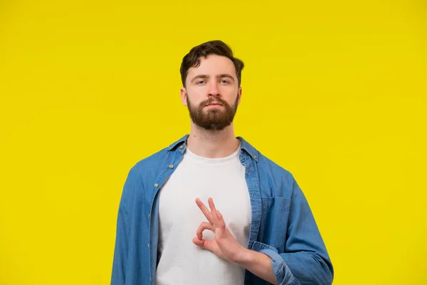 Portrait of handsome excited man in basic clothing smiling and showing ok sign at camera isolated over yellow background